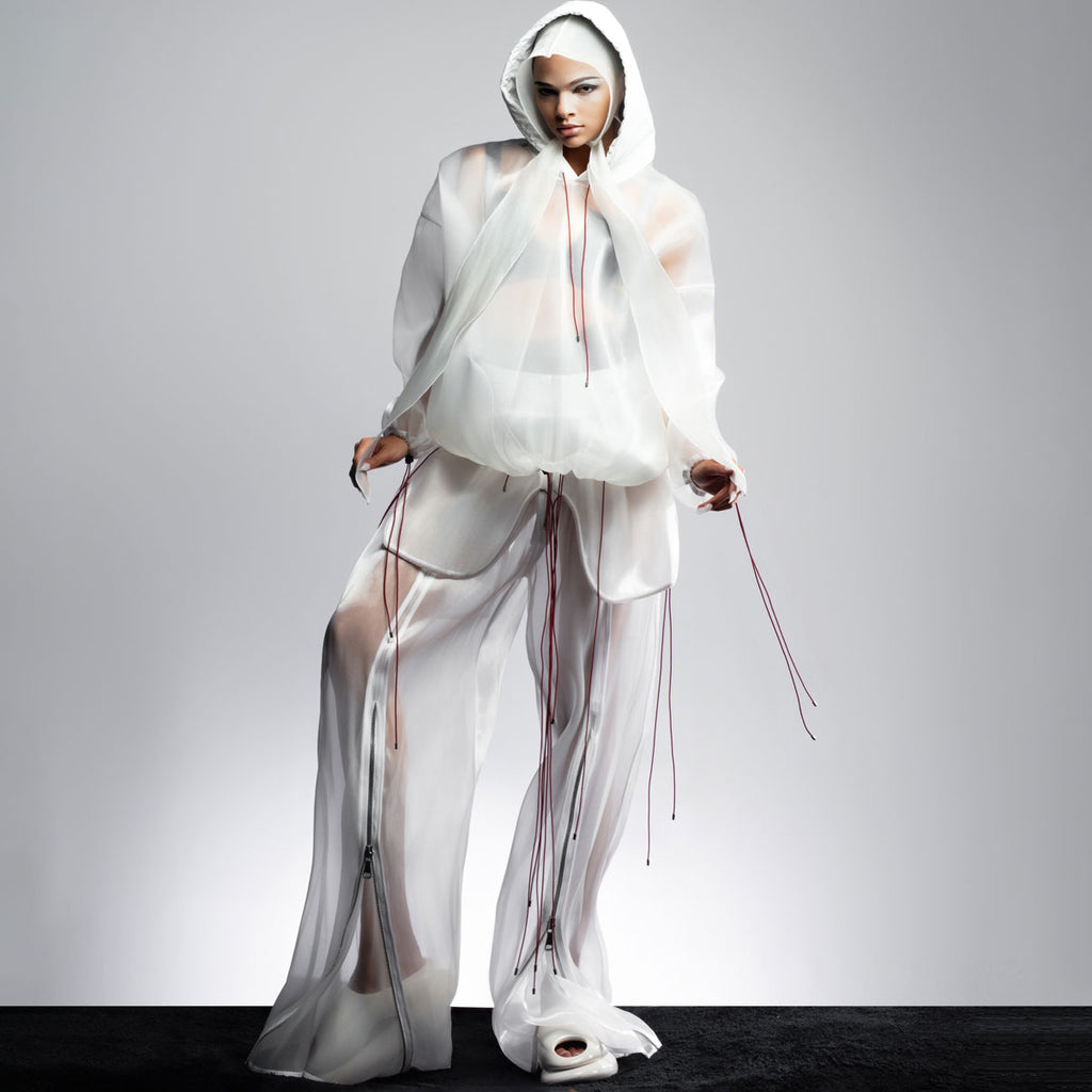Model wearing a white sheer tracksuit outfit, a jacket and pants both oversized with red drawstrings and zips.